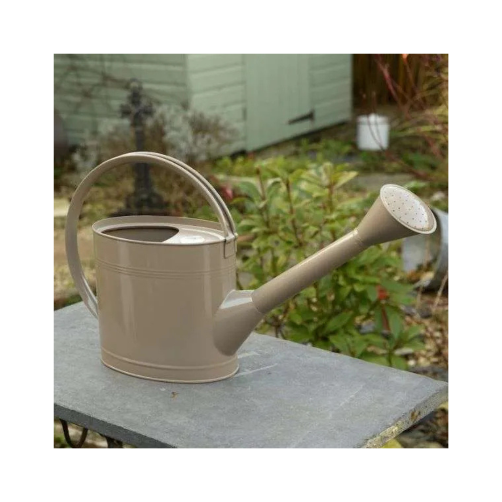 Truffle-coloured 5L metal watering can placed on a garden table with greenery and a shed in the background, showcasing its long spout and ergonomic handle.